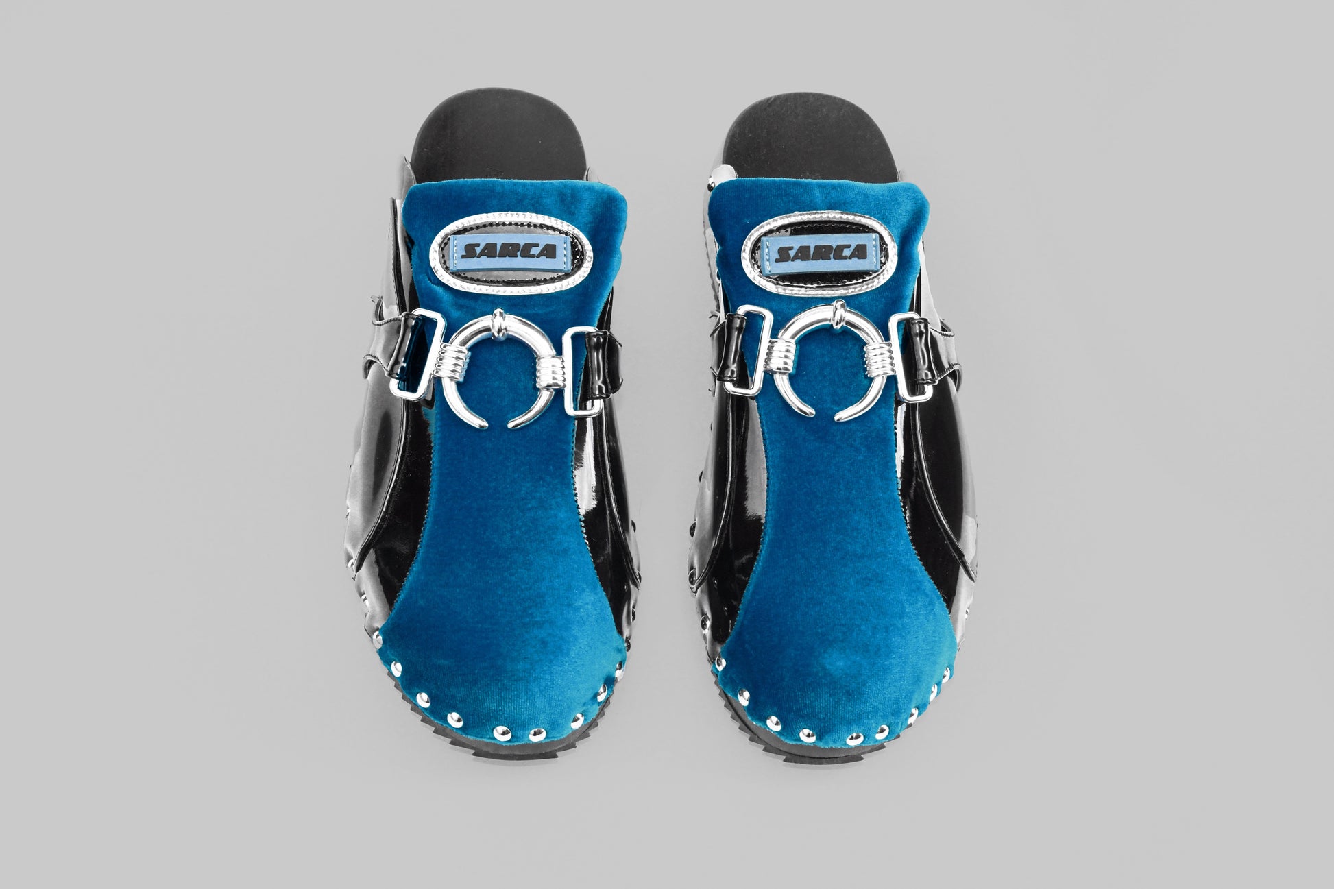 A photograph of the top view of a pair of Ancuta Sarca blue back beetle clogs, patent finish, velvet panel with ring embellishment, logo patch at tongues, branded footbeds, round toes, silver tone hardware, made in Italy. Unisex shoe.